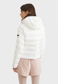Veste thermique blanche avec capuche, design matelassé, texture lisse et ajustement réglable. Présente une accent noir sur la manche.