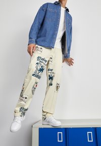 Camicia di denim con bottoni davanti, abbinata a pantaloni beige con audaci stampe grafiche blu. Sneakers bianche completano l'outfit.