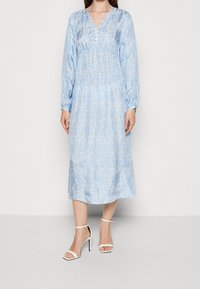 Vestido midi floral azul claro con mangas largas, escote en V, cintura elástica y detalles de botones. Llevado con tacones blancos de tira.