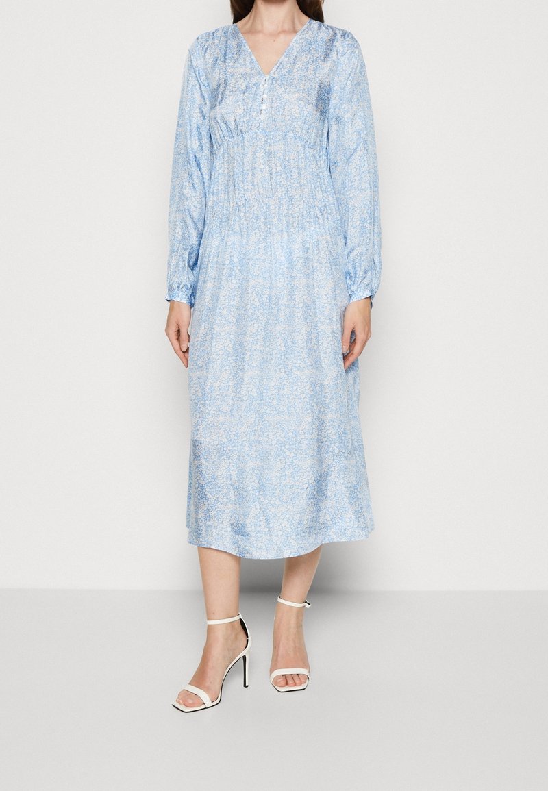 Vaaleansininen kukkakuosis midi-mekko pitkillä hihnoilla, V-pääntiellä, elastisella vyötäröllä ja napitusyksityiskohdalla. Yhdistetty valkoisten hihnakenkien kanssa.