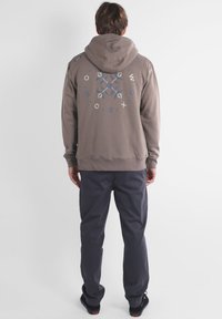 Sweatshirt à capuche gris avec un imprimé géométrique bleu et crème au dos. Associé à un pantalon foncé et des chaussures noires. Texture plate.