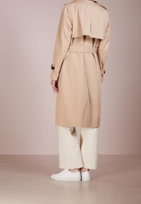 Cappotto beige con vita cintrata, ampio colletto e dettaglio della ventaglietta posteriore, abbinato a pantaloni bianchi a gamba larga e sneakers bianche.