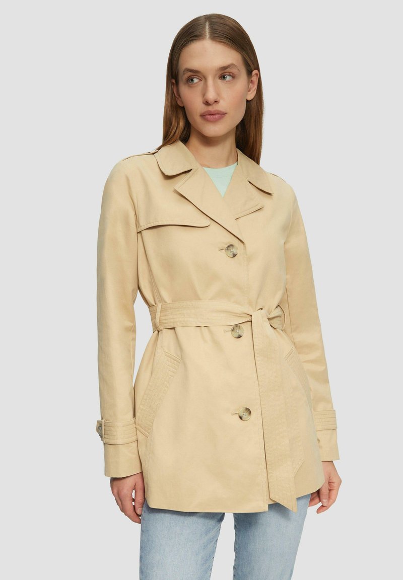 Beige trench coat s páskem v pase, velkými klopami a čtyřmi knoflíky. Obsahuje boční kapsy a texturované detaily na rukávech.