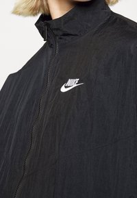 Nike svart jacka med hög krage, texturerad tyg, framsidan med dragkedja och en liten vit logotyp på vänster bröst. Enkel design, lättviktig.
