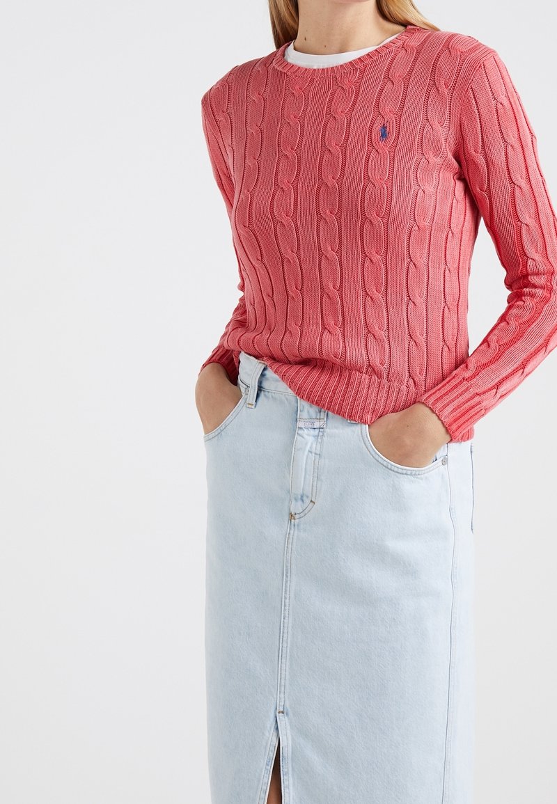 Femme portant un pull torsadé rose et une jupe en denim bleu clair avec une fente devant, mains dans les poches de la jupe, sur fond blanc.
