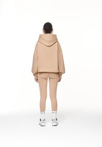 Mujer de pie, de espaldas, con sudadera beige, leggings a juego, calcetines blancos y zapatillas blancas, sobre fondo blanco.