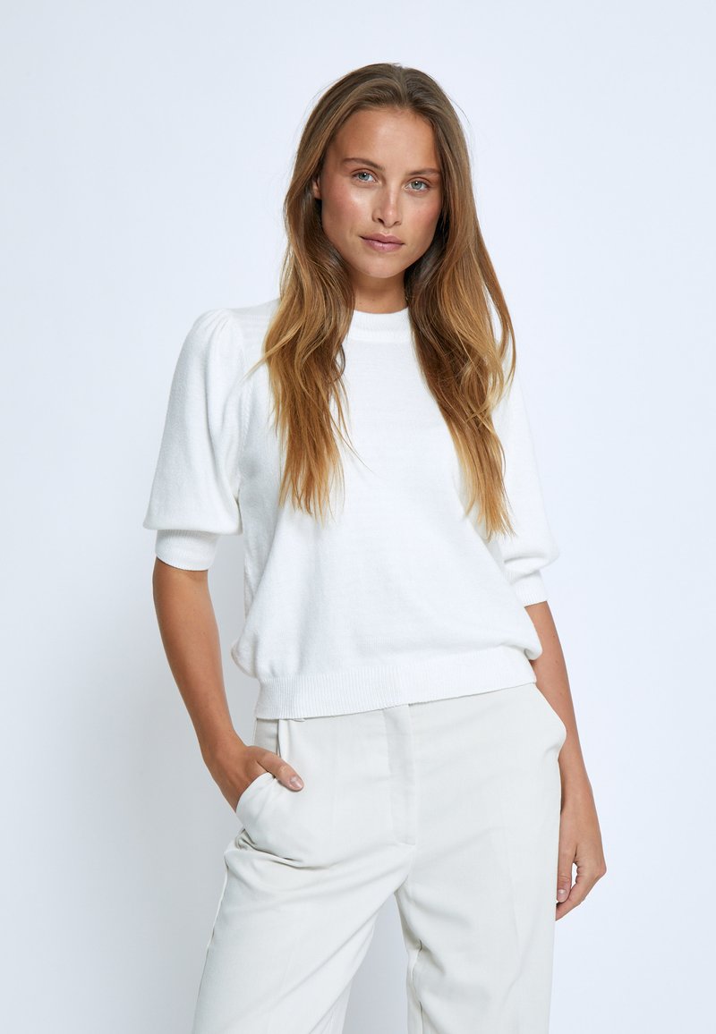 Weißer gestrickter Pullover mit kurzen Ärmeln, Rundhalsausschnitt und geripptem Saum. Weiche Textur, leicht puffige Ärmel, kombiniert mit hellfarbigen Hosen.