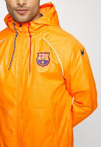 Orange vindjacka med reflekterande detaljer, med huva, dragkedjestängning och en framträdande FCB-logotyp på bröstet. Strukturerad tyg med mönster.