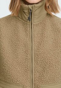 Beige Fleecejacke aus strukturiertem, flauschigem Material und einem kontrastierenden glatten Stoff im Reißverschlussbereich. Mit hohem Kragen und Fronttaschen.