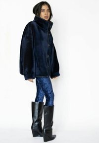 Marineblå faux fur-jakke med høj krave og knappelukning foran, kombineret med glitrende blå fløjelsskindsleggings og knæhøje sorte kilehælstøvler.