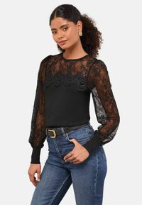 Fekete hosszú ujjú felső, áttetsző csipke bodiccal és ujjal, részletes csipke díszítéssel és egy sima fekete, testhez simuló alsó résszel. Kék farmerral párosítva.