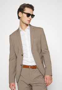 Light brown tailored suit with a white shirt featuring small blue patterns. Brown belt, green-tinted sunglasses, and structured design.