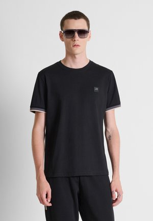 Jonge man die een zwart T-shirt met logopatch en zwarte zonnebril draagt, staand tegen een effen lichtgrijze achtergrond.