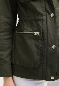 Kurtka oliwkowozielona wykonana z miękkiego, teksturowanego materiału, z kieszenią na suwak z srebrnym okuciem oraz detalami na przodzie w postaci zatrzasków.