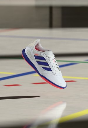 Bílá indoorová fotbalová bota s modrými pruhy a červenou podrážkou vznášející se nad podlahou tělocvičny označenou barevnými čarami.