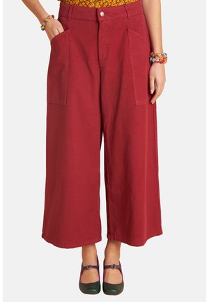 Frau trägt weit geschnittene, gekürzte rote Hosen mit großen aufgesetzten Taschen vorne, grüne Mary-Jane-Schuhe und bunte Perlenarmbänder an beiden Handgelenken.