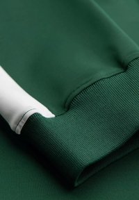 Grön tröja med vit accent; ribbade ärmslut; slitstark yta; klassisk passform; materialet verkar lätt och ventilerande.