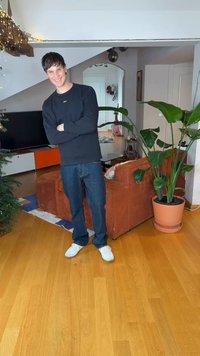 Νέος άνδρας με μαύρο φούτερ και μπλε τζιν βρίσκεται σε ένα σαλόνι με καφέ καναπέ από κοτλέ, μεγάλο γλαστράκι και ξύλινο πάτωμα.