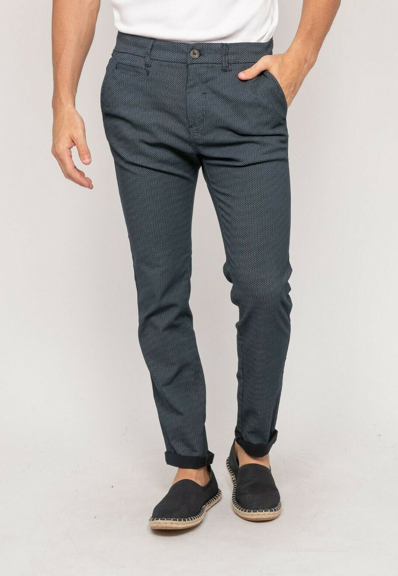 Pantalons texturés bleu marine avec une coupe slim, dotés d'une fermeture à bouton et de poignets retroussés, associés à des chaussures de style espadrilles foncées.