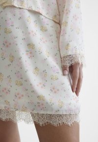 Blomstret kjole med en lys kremfarget stoff med rosa, gule og grønne blomster mønstre. Blondekantene ved hem og ermer gir tekstur.