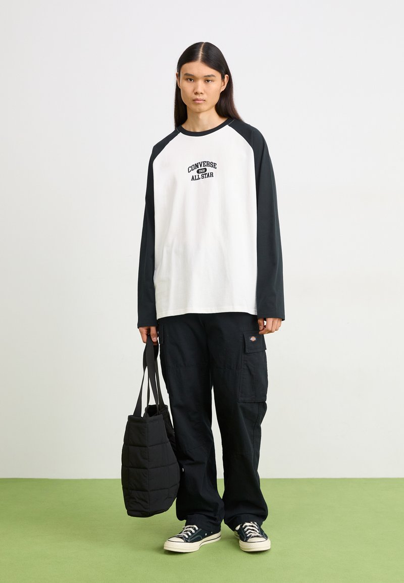 Chemise blanche à manches longues avec manches raglan noires, présentant un graphique "Converse All Star". Associée à un pantalon cargo noir et un sac noir matelassé.