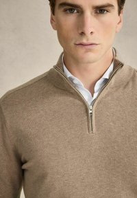 Beige Zip-up-Pullover aus weichem Stoff, mit hohem Kragen und langen Ärmeln, über einem weißen Hemd mit Knopfleiste getragen.