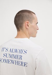 Balta medvilninė marškinė su mėlyna tekstu ant nugaros, rašančiu "VISADA YRA VASARA KUR NORS." Apvalus kaklas, trumpomis rankovėmis.