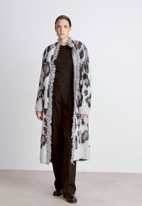 Cardigan lungo con frange in materiale lavorato a maglia, caratterizzato da un motivo leopardato grigio e beige. Indossato sopra una maglietta nera e pantaloni neri.