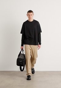 Black short-sleeve T-shirt with embossed logo, paired with beige wide-leg trousers. The model carries a black duffel bag and wears black shoes.