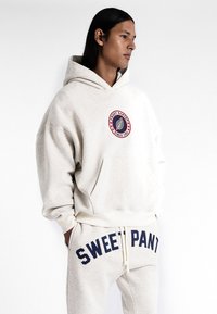 Haut à capuche gris clair avec un grand logo à l'avant et un texte bleu foncé portant l'inscription « SWEET PANTS ». Pantalons de survêtement gris clair coordonnés avec un texte assorti.