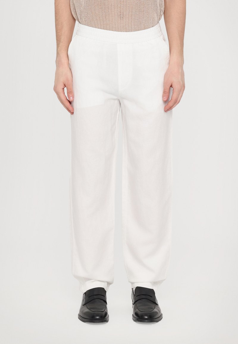 Pantalons en lin blancs avec une taille élastique, offrant une coupe décontractée et des poches latérales, associés à des chaussures noires.