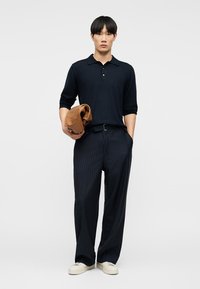 Άνδρας με μαύρο πόλο με μακριά μανίκια, μαύρο παντελόνι με ψιλή ρίγα, λευκά αθλητικά παπούτσια, κρατώντας ένα διπλωμένο καφέ σακάκι στο αριστερό χέρι.