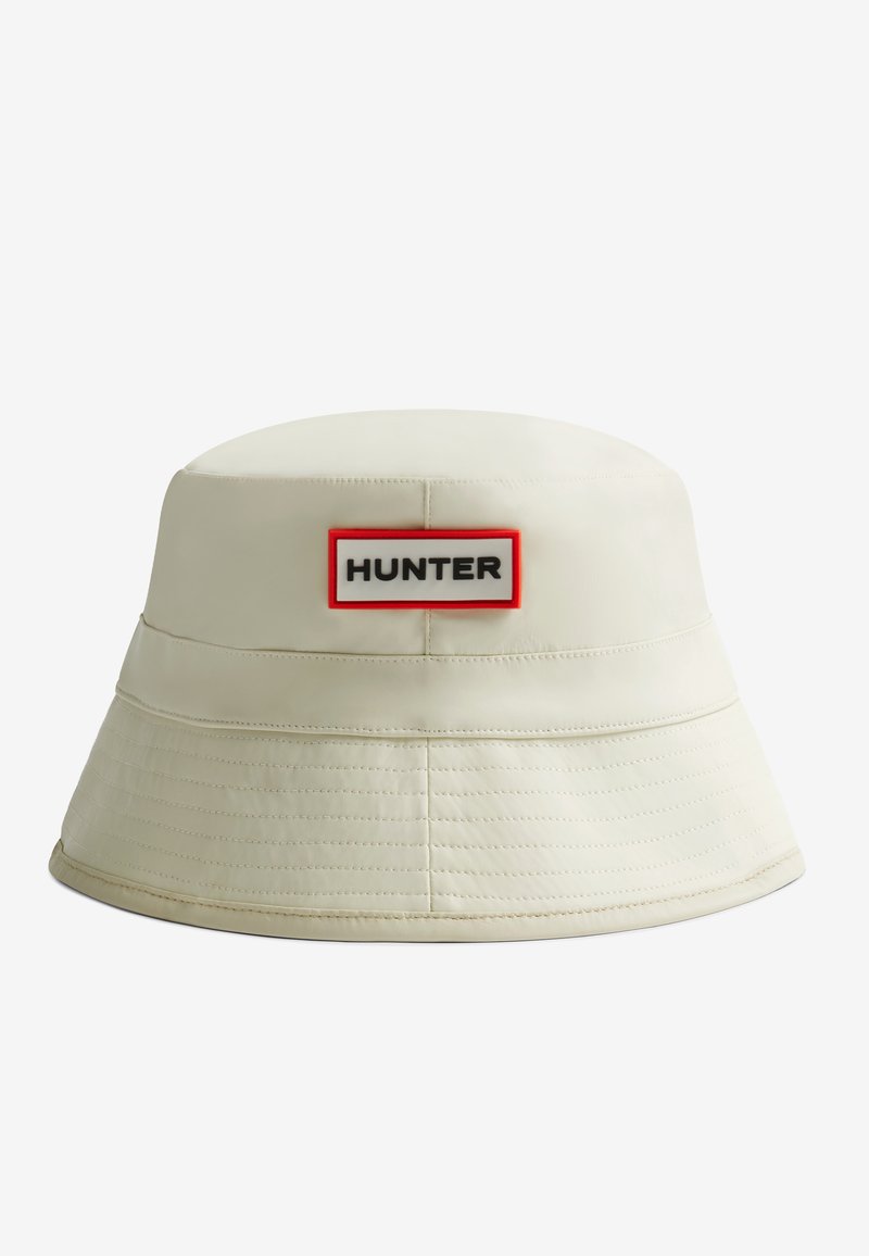 Krémový vedro klobúk vyrobený z hladkého materiálu, s okrúhlym vrchom a širokým krempou. Na ňom je viditeľná vyšívaná etiketa "HUNTER" v červenom okraji.
