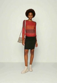 Mulher em pé, vestindo uma blusa de mangas compridas em gradiente vermelho, saia preta até o joelho, botas bege e segurando uma mala de ombro bege.