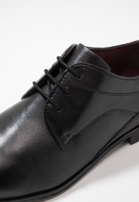 Fekete bőrből készült elegáns cipő sima felülettel, fűzős orral és diszkrét vonalvezetéssel. Jellemzője egy varrott varrás és egy bordó belső rész.