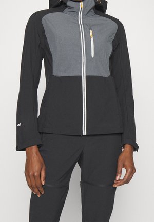 Femme portant une veste à capuche zippée noire et grise avec une poche poitrine et un pantalon noir assorti, debout devant un fond blanc.