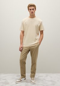 T-shirt beige a maniche corte con scollo rotondo, abbinata a pantaloni slim-fit beige chiaro e sneakers beige con dettagli bianchi.