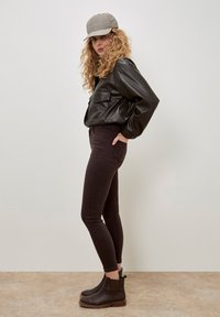 Chaqueta de cuero negra corta, jeans ajustados marrón oscuro y botas negras hasta el tobillo con suela marrón. Se lleva una gorra de espiga gris.