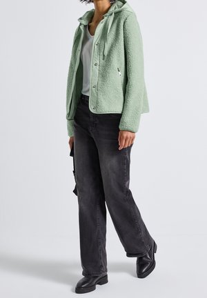 Persoon die een lichtgroene fleecejas, wit overhemd, zwarte wijde jeans en zwarte laarzen draagt, staand tegen een effen achtergrond.