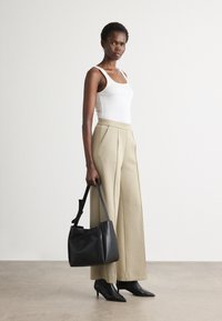 Beige hoge taille broek met plooien, gecombineerd met een witte tanktop. Houdt een zwarte leren schoudertas vast en draagt spitse zwarte hakken.