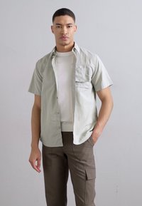 Lys grøn kortærmet skjorte med en lomme, lagdelt over en hvid t-shirt. Sammen med brune cargo-bukser med sidelommer.