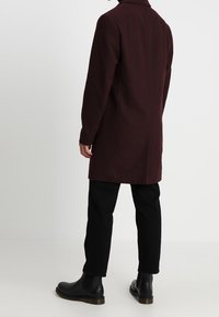 Cappotto in misto lana bordeaux con design diritto, colletto tagliato e maniche lunghe. Abbinato a pantaloni neri e stivaletti neri.