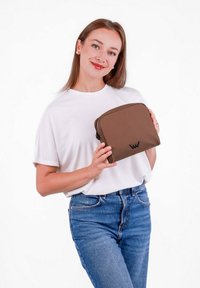 Braune kosmetische Tasche aus Stoff in rechteckiger Form, mit glatter Textur und einem schwarzen Logo auf der Vorderseite, gehalten von einer Person in einem weißen Hemd.