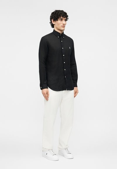 Hombre con camisa negra de manga larga abotonada, pantalones blancos y zapatillas blancas, de pie contra un fondo claro y liso.