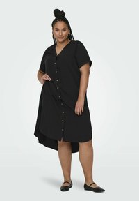 Robe noire à boutons, avec un décolleté en V et des manches courtes. Dotée de fentes sur les côtés et d'un ourlet arrière légèrement plus long. Fabriquée en tissu doux.