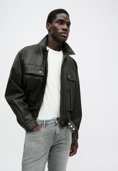 Μαύρο δερμάτινο μπουφάν με ψηλό γιακά, ασημένιες λεπτομέρειες με χρυσά κουμπιά και πολλές μπροστινές τσέπες, συνδυασμένο με λευκό μπλουζάκι και γκρι τζιν.