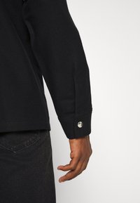 Bras droit d'une personne portant une chemise noire à manches longues avec un bouton-pression en métal sur le poignet et un jean en denim foncé sur fond blanc.