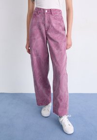 Jeans taille haute acid wash rose clair avec une coupe décontractée, dotés de cinq poches et de baskets blanches, sur un sol bleu.