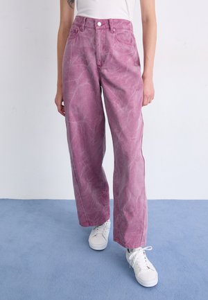 Light pink acid-wash high-waisted jeans with a relaxed fit, featuring five pockets and white sneakers, set against a blue floor.