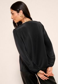 Black satin blouse with long sleeves and gathered cuffs, featuring a smooth texture and a rounded neckline, worn with black pants.
